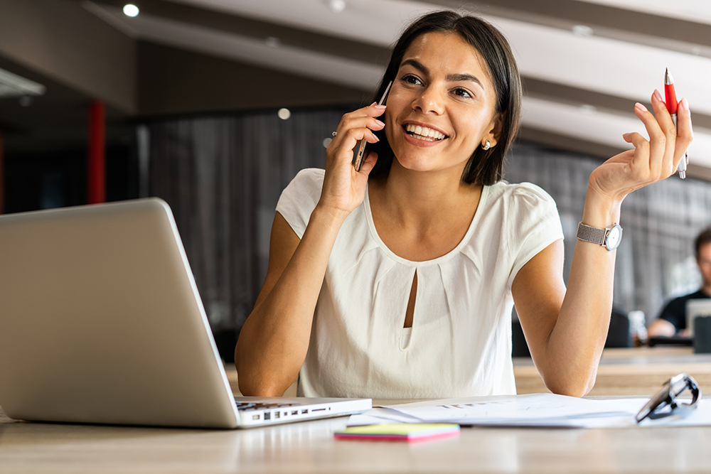 Cheerful young woman talking on mobile phone and using laptop with smile while sitting at her working place
