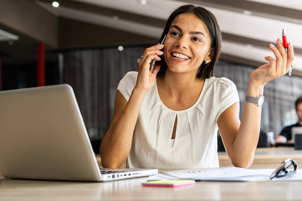 Lady sitting on desk with laptop while talking on phone with pen in hand